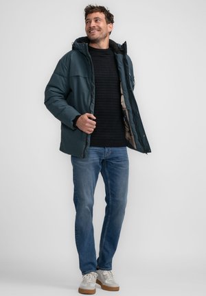 Chaqueta acolchada en azul oscuro con capucha, llevada sobre un suéter negro de canalé, combinada con jeans azules claros y zapatillas grises con suelas marrones.