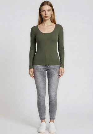 Jonge vrouw die recht vooruit kijkt, draagt een olijfgroene top met lange mouwen, grijze acid-wash skinny jeans en witte sneakers.