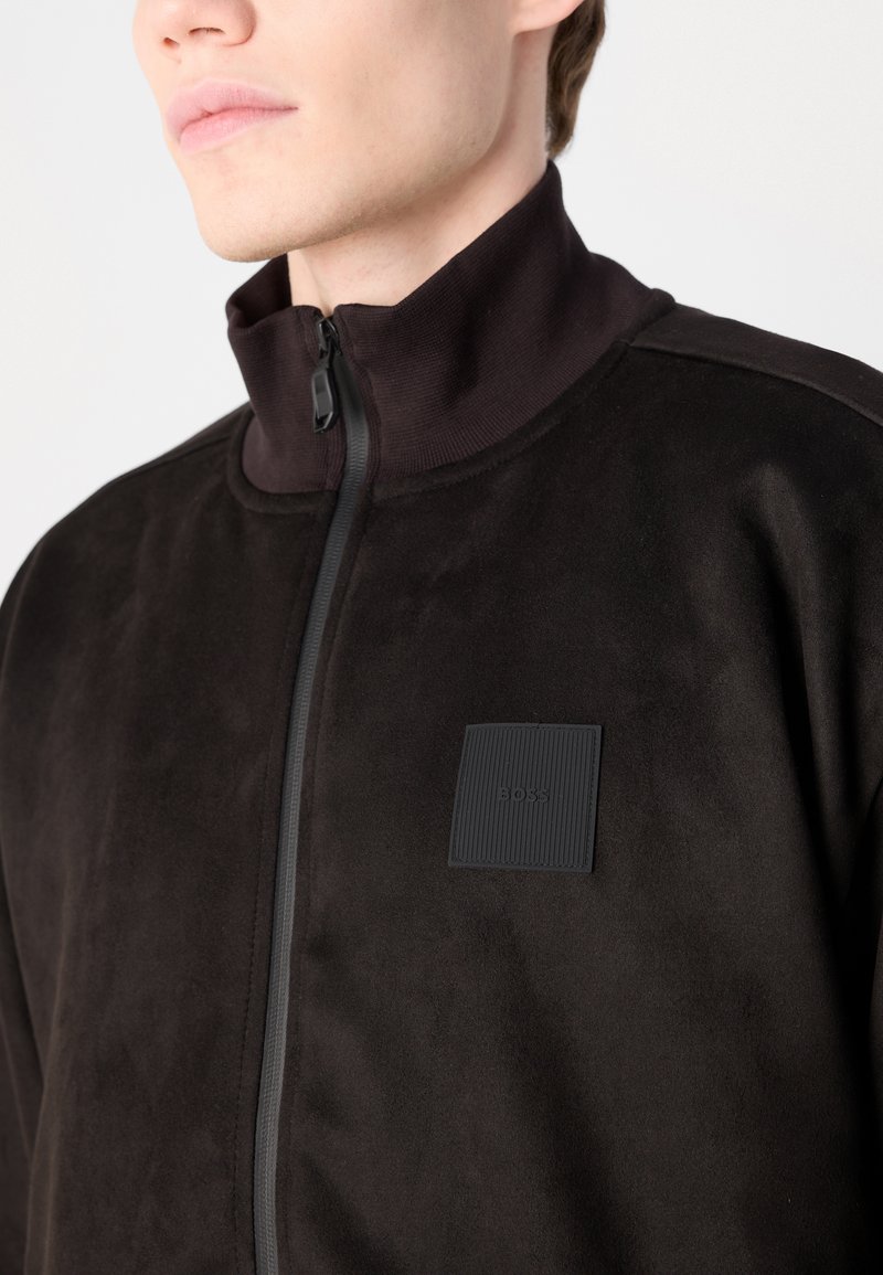 Schwarze Zip-Jacke aus weichem Stoff mit hohem Kragen, ausgestattet mit einem strukturierten quadratischen Logo-Patch und einer glatten Oberfläche.
