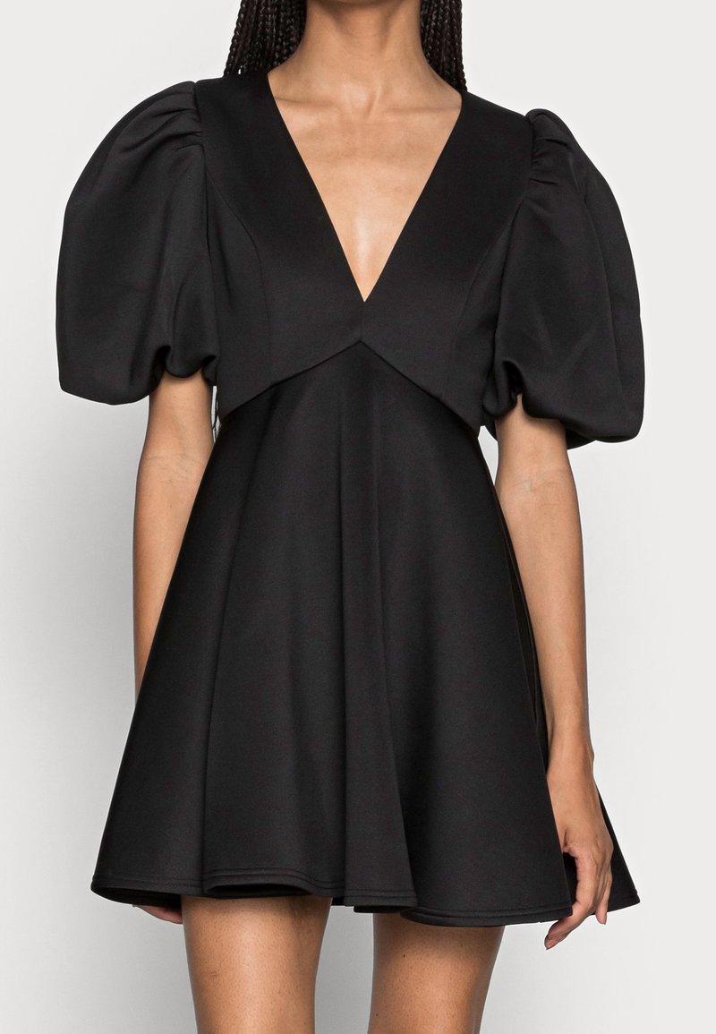 Femme portant une robe courte noire avec un profond décolleté en V et des manches bouffantes volumineuses, debout devant un fond uni.