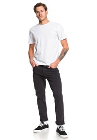 T-shirt en coton blanc, pantalon noir et baskets noires avec des semelles blanches ; coupe décontractée avec les ourlets de pantalon retroussés ; tatouage sur le bras.