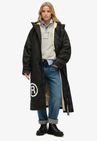 Abrigo largo negro con logo blanco, frente con cremallera y cuello forrado de piel. Lleva puesto sobre un sudadera gris, una camisa azul y unos vaqueros con puños enrollados, junto con botas.