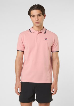 Junger Mann mit kurzen braunen Haaren, der ein rosa Poloshirt mit schwarzem Rand und schwarze Shorts trägt, stehend vor einem schlichten hellen Hintergrund.