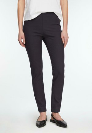 Donna che indossa pantaloni neri aderenti, top bianco e ballerine nere con cinturino, in piedi su sfondo bianco.