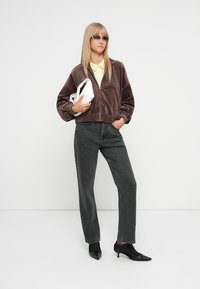 Smeđa, plišana jakna na zatvaranje s elastičnim manžetama, u kombinaciji s crnim hlačama ravnog kroja. Bijela torbica i žuta košulja s kragnom upotpunjuju izgled.