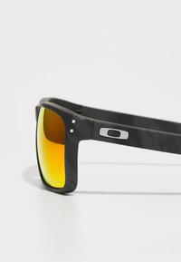Ochelari de soare din plastic negru, cu o ramă pătrată, având lentile de culoare portocalie și accente argintii pe sprâncene. Textură mată.