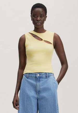 Woman wearing a sleeveless pale yellow top with cutouts and high-waisted blue jeans, standing with one hand in pocket against plain background.
