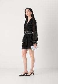 Black pleated dress with long sleeves, tiered design, silver belt accent, and open-toe black slingback heels. Silver clutch in hand.
