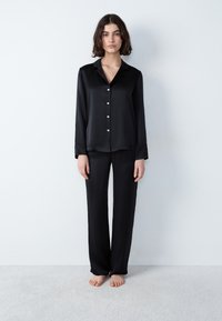 Conjunto de pijama de satén negro que incluye una camisa de botones de manga larga con cuello y pantalones holgados a juego. Textura suave, con detalles mínimos.