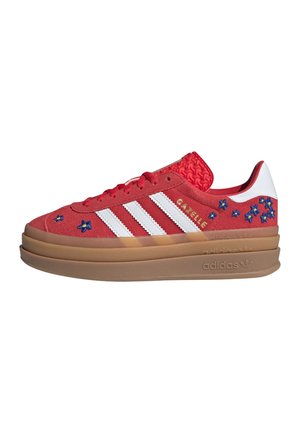 Czerwony sneaker Adidas Gazelle z białymi paskami, niebieską kwiatową haftą, wytłacznym czerwonym językiem i grubą brązową podeszwą.