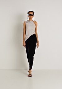 Aszimmetrikus bézs felső fekete bársony nadrággal párosítva, amely sima textúrával és oldalsó hasítékkal rendelkezik. A modellen napszemüveg és pántos fekete magassarkú cipők találhatók.
