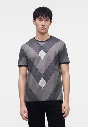 Giovane uomo che indossa una camicia grigia e nera a maniche corte con un motivo geometrico a chevron e diamante, in piedi davanti a uno sfondo chiaro e uniforme.