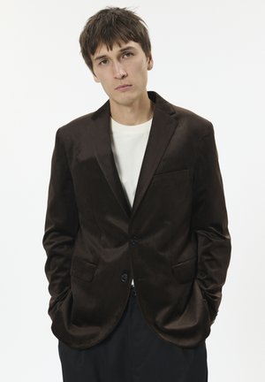 Brauner Samtblazer mit Reverskragen, zwei vorderen Taschen und zwei Knöpfen, kombiniert mit einem weißen Rundhalshemd darunter.