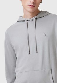 Sudadera gris claro con cordones ajustables, bolsillo tipo canguro en la parte frontal y un pequeño logo bordado en el pecho. Textura de tela suave.