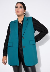 Blazer sin mangas turquesa con dos bolsillos frontales y dos botones, llevado sobre una camisa negra a rayas. Tela suave y estructurada.