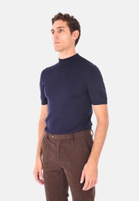 Uomo in piedi, indossa una maglietta a maniche corte con collo alla coreana blu navy e pantaloni di velluto a coste marroni, rivolto leggermente verso sinistra di fronte a uno sfondo neutro.