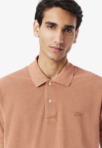 Ljusbrun polo skjorta med en texturerad tyg, med krage och tre knappar fram. Lacoste-logotypen broderad på vänster bröst.