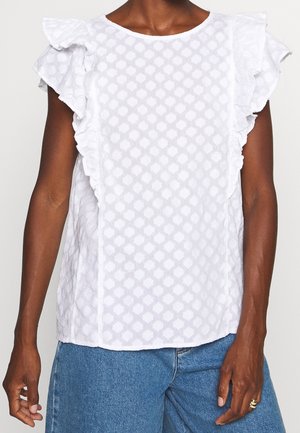 Blusa blanca sin mangas con hombros con volantes y un sutil patrón geométrico, usada con vaqueros azules por una persona de piel oscura.