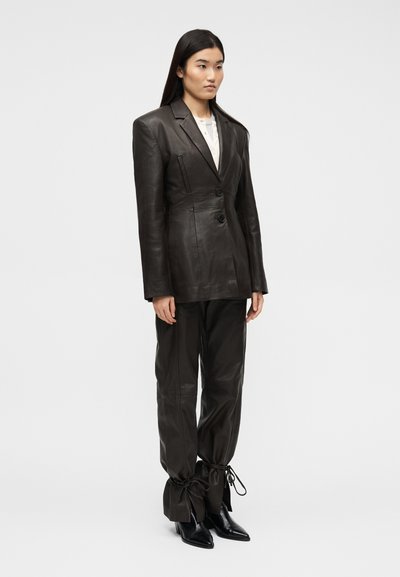 Femme portant un blazer et un pantalon en cuir marron foncé avec les chevilles nouées, assortis à des bottes noires à bouts pointus, debout devant un fond uni.