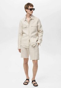 Lanena srajca v svetli bež barvi z dvema sprednjima žepoma in gumbom, kombinirana z ustreznimi kratkimi hlačami in črnimi sandalami. Mat finiš, sproščen kroj.