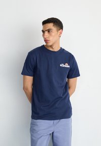 T-shirt en coton bleu marine avec un col rond et des manches courtes, arborant un logo blanc sur la poitrine. Porté avec un pantalon violet clair.