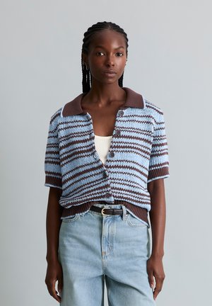 Frau mit geflochtenem Haar, die eine blau-braun gestreifte kurzärmelige Strickjacke, ein weißes Oberteil, hellblaue Jeans und einen schwarzen Gürtel trägt, steht vor einem grauen Hintergrund.