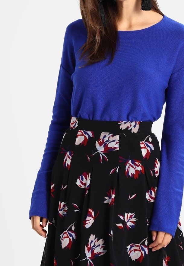 Femme portant un haut bleu roi à manches longues, rentré dans une jupe noire taille haute avec des motifs floraux rouges, blancs et violets.