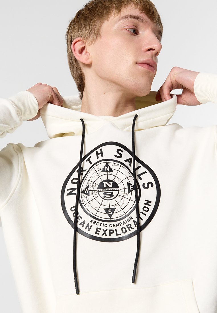 Joven ajustando una sudadera con capucha crema con un diseño de brújula negra y el texto "North Sails Arctic Campaign Ocean Exploration" en el frente.
