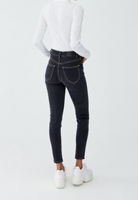 Vysoké černé džíny s úzkým střihem, vyrobené z texturované látky, s kontrastním prošíváním a zadními kapsami. Stylizované s bílými teniskami.