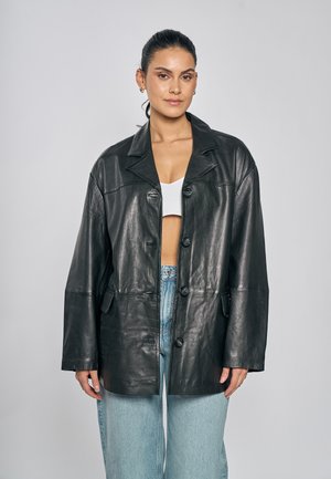 Kvinde med mørkt hår sat op, iført en sort, oversized læderjakke, hvid crop top og lyseblå jeans mod en ensfarvet baggrund.