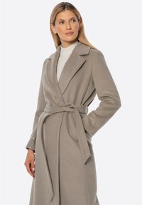 Světle šedý vlněný kabát s páskem, dlouhými rukávy a klopovým límcem. Disponuje knoflíkovými detaily na manžetách pro přidanou eleganci a strukturu.