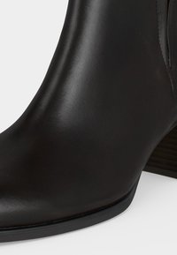 Gros plan d'une botte cheville en cuir noir lisse avec un talon bas empilé et un bout pointu sur fond blanc.