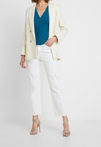 Blazer beige à double boutonnage sur un haut teal, associé à un pantalon droit blanc et des sandales à talons argentées avec des lanières entrecroisées.