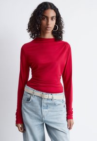 Rotes Langarmoberteil aus glattem Stoff mit einem drapierten Design. Kombiniert mit hellblauen, hochgeschnittenen Jeans und einem hellen Gürtel.