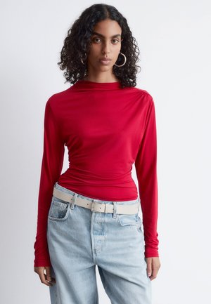 Rotes Langarmoberteil aus glattem Stoff mit einem drapierten Design. Kombiniert mit hellblauen, hochgeschnittenen Jeans und einem hellen Gürtel.