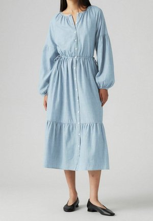 Vrouw draagt een lichtblauwe, lange mouwen, knoopjes midi-jurk met elastische tailleband en zwarte lage hakken, staand op een grijze achtergrond.