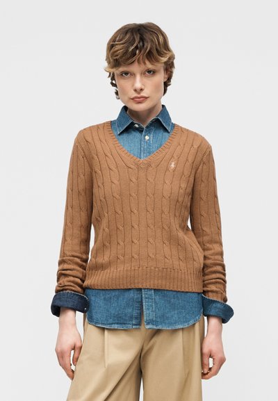 Άτομο που φοράει καφέ πλεκτό πουλόβερ με V λαιμόκοψη πάνω από μπλε τζιν πουκάμισο με γυρισμένα μανίκια και μπεζ παντελόνι, με φόντο απλό.