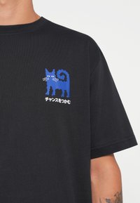 Zwarte t-shirt met een blauwe afbeelding van een kat met opvallende snorharen, een gekrulde staart en Japanse tekst onder het ontwerp.