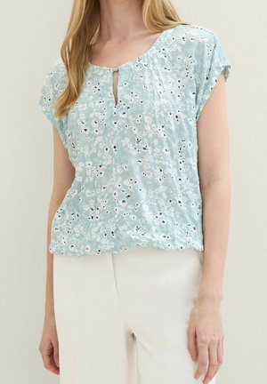 Mujer con blusa azul claro de manga corta con flores y escote en forma de ojo de cerradura, y pantalones blancos, de pie frente a un fondo liso y claro.