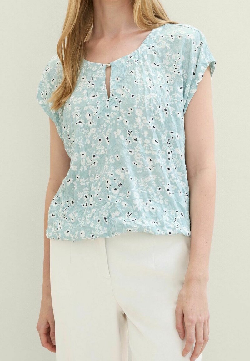 Femme portant un chemisier bleu clair à manches courtes fleuri avec un décolleté en forme de goutte et un pantalon blanc, debout devant un fond clair uni.