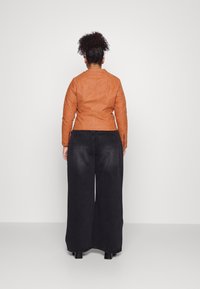 Giacca in finta suede color ruggine con un design aderente, abbinata a jeans di denim neri larghi. Entrambi gli articoli presentano dettagli minimi.