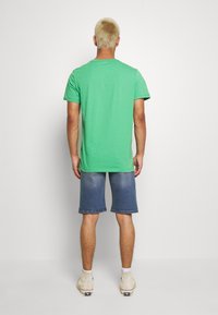 Hombre con cabello corto rubio que lleva una camiseta verde de manga corta, pantalones cortos de mezclilla azul, calcetines blancos y zapatillas altas beige, de pie de espaldas.