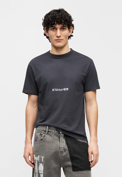 Σκούρο γκρι μπλουζάκι με το λογότυπο "KIDSUPER" σε λευκό, κοντά μανίκια, στρογγυλή λαιμόκοψη, συνδυασμένο με γκρι τζιν που εμφανίζει διάφορες υφές και μπαλώματα.