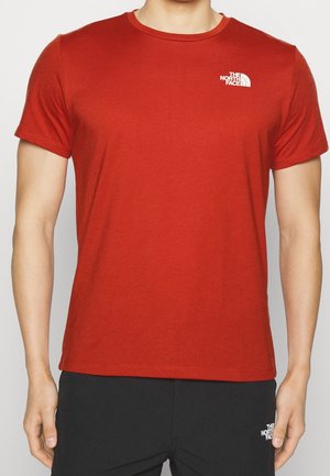 Mies, joka pukeutunut yksiväriseen punaiseen lyhythihaiseen T-paitaan, jonka vasemman rinnan yläosassa on valkoinen The North Face -logo.