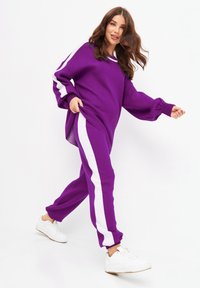 Pull en maille oversize violet avec des rayures blanches sur les manches, associé à un pantalon violet assorti avec des rayures blanches sur les côtés. Baskets blanches.
