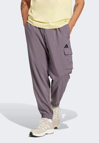 Pantalones cargo en un púrpura claro, fabricados con un material ligero. Presentan puños elásticos, un bolsillo lateral y acentos del logo negro de Adidas.