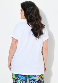 T-shirt blanc à manches courtes avec des ourlets retroussés, coupe décontractée, texture lisse, porté avec des leggings colorés à motifs présentant des designs graphiques.