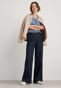 Blusa a fiori blu, giacca oversize beige, jeans a gamba larga blu scuro e scarpe da ginnastica chiare. Outfit semplice e casual.