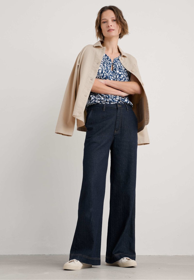Blusa a fiori blu, giacca oversize beige, jeans a gamba larga blu scuro e scarpe da ginnastica chiare. Outfit semplice e casual.