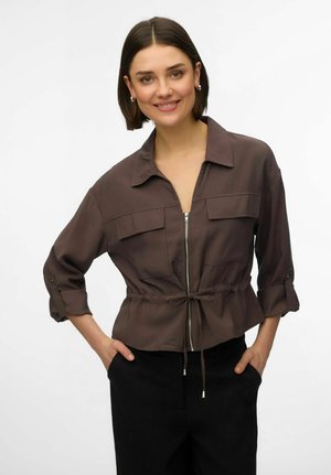 Femme aux cheveux courts bruns souriante, portant une veste marron zippée aux manches retroussées et un pantalon noir, les mains dans les poches.